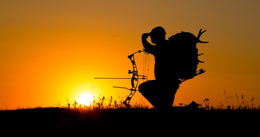 Man holding bow and hunting gear with sunset in background